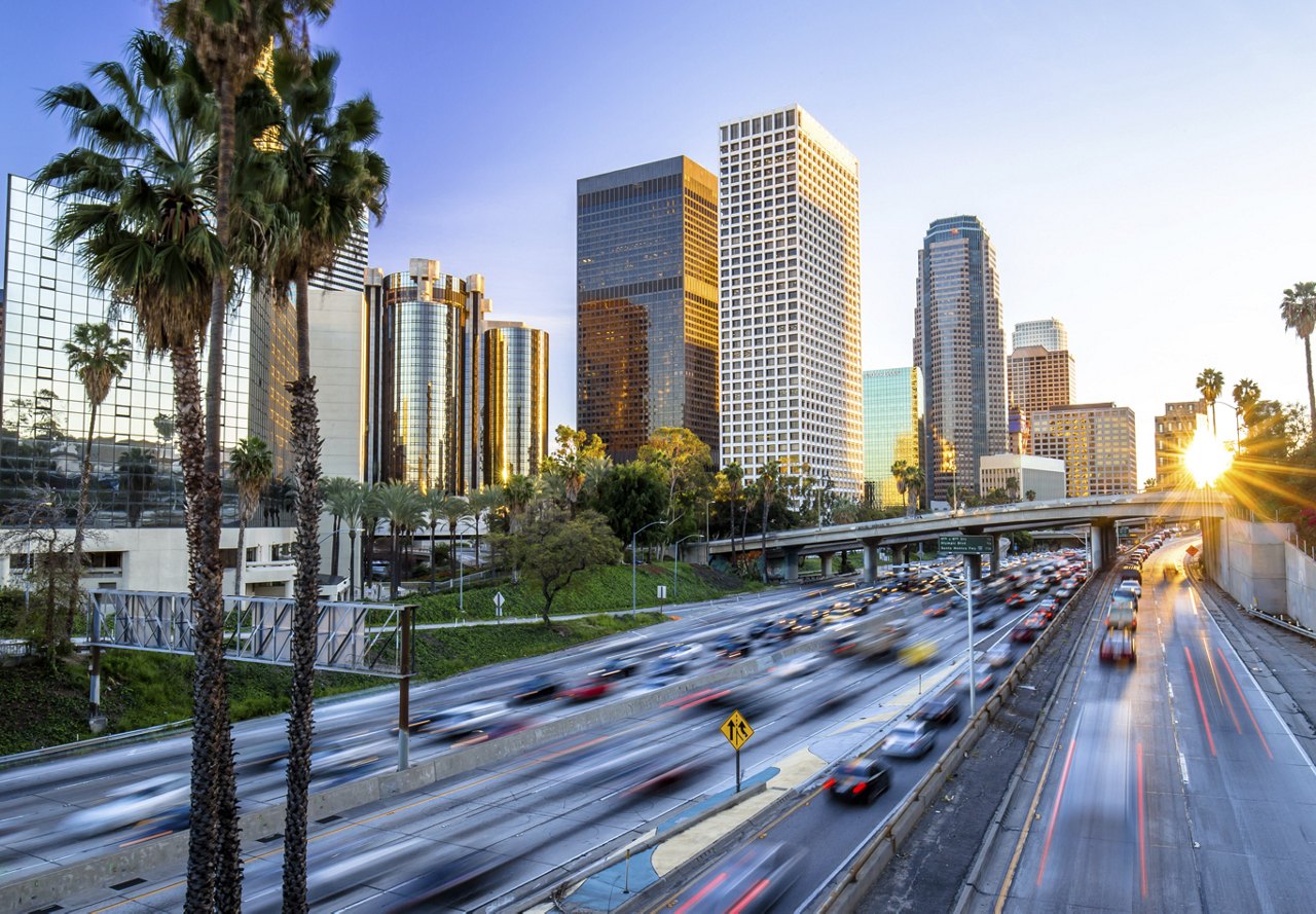 Busy interstate in downtown Los Angeles, with cars zooming by, tall skyscrapers in the background, palm trees lining the road, and a sunset lighting up the scene.