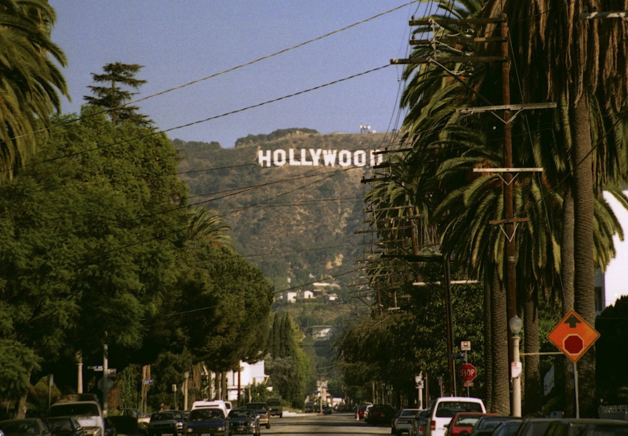 View of the Hollywood sign in the distance, with trees lining the street and cars parked along the side of the road.