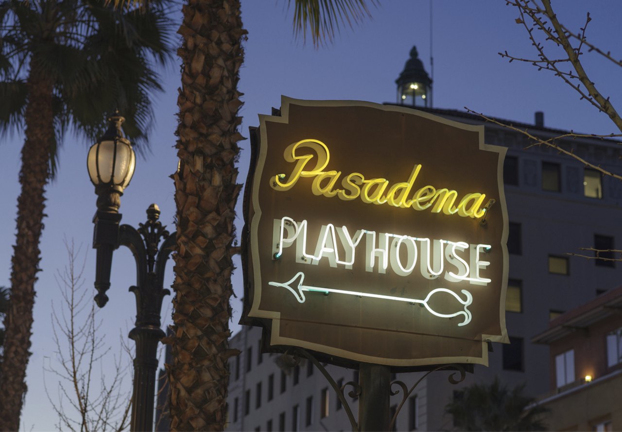 Pasadena Playhouse sign illuminated in neon at night, surrounded by trees and buildings.