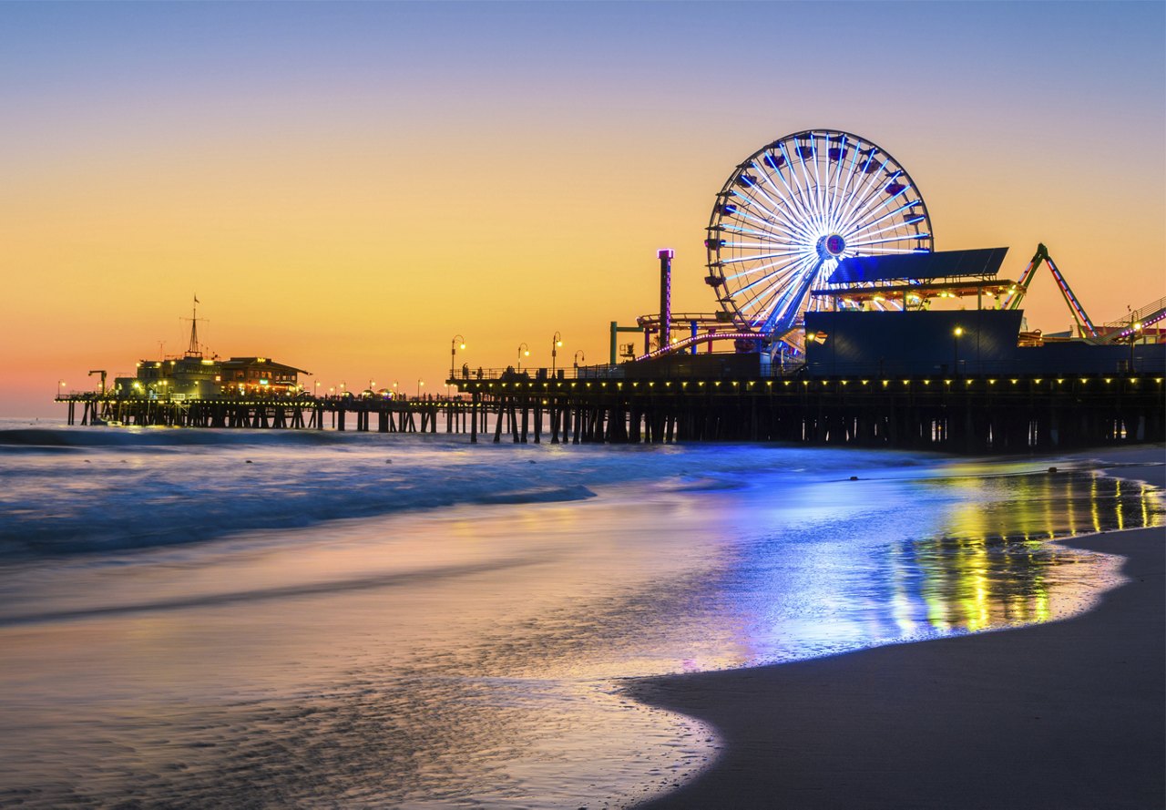 Beautiful sunset view of the Santa Monica Pier, featuring a glowing Ferris wheel and vibrant lights across the pier, with the ocean in the foreground.