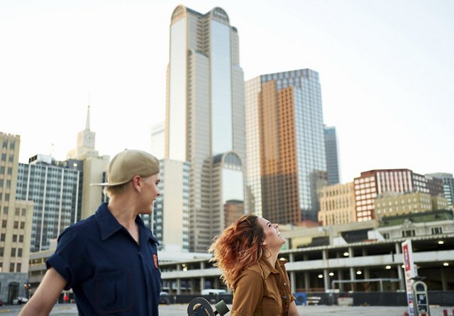 Two young people skateboarding through Dallas, gazing up at the city's towering buildings.