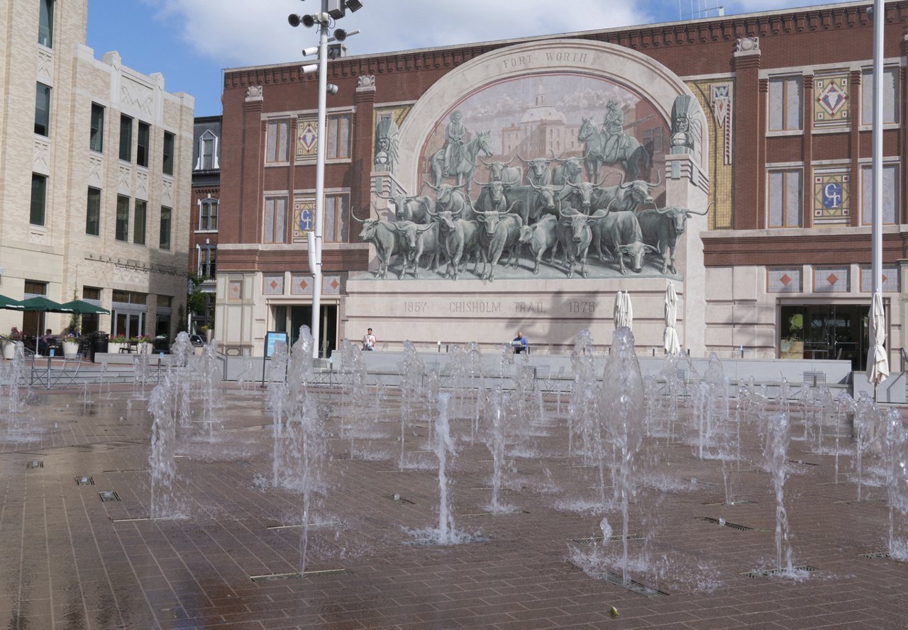 Fountains spouting from the ground in Fort Worth, with a historic sign in the background, surrounded by brick architecture.