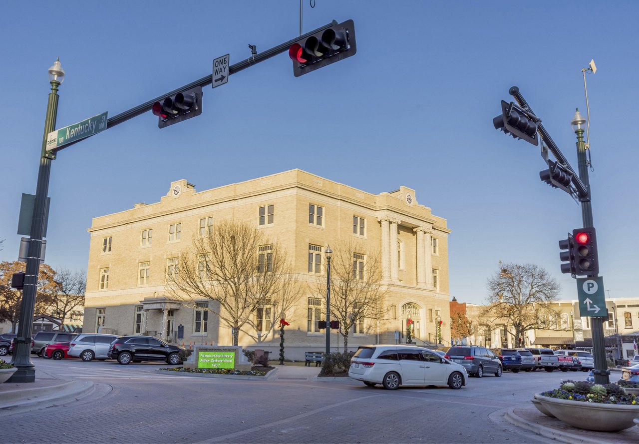 Downtown McKinney, Texas, showing cars passing through a lighted intersection with a government building in the background during the day.