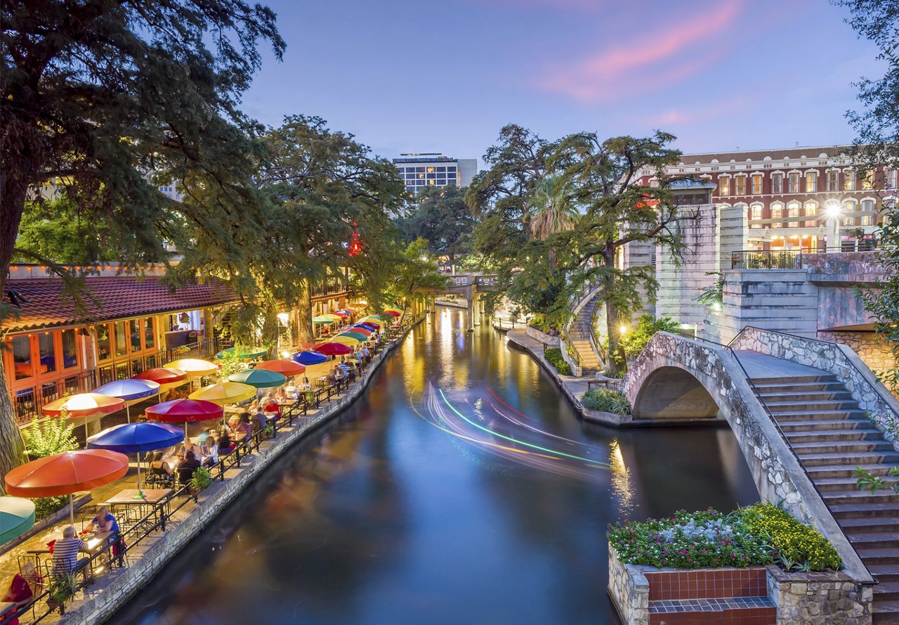 Nighttime view of the San Antonio River Walk, illuminated with numerous lights and umbrellas along the canal.