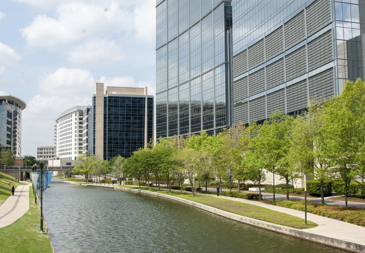 View of a waterway running through The Woodlands, Texas, surrounded by tall buildings, trees, and walking pathways on either side.