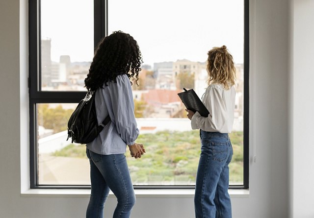 Two women standing by an apartment window, one holding a clipboard, overlooking the cityscape.