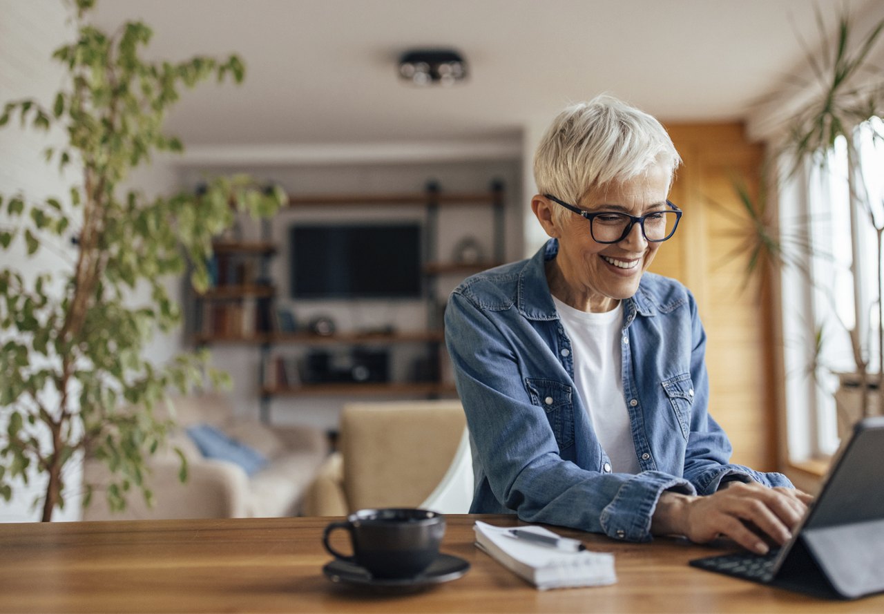 Older woman smiling while typing on a computer in her apartment, with a coffee cup nearby and surrounded by lush plants.