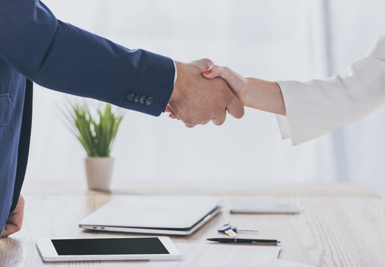 Two people shaking hands in an office setting, with a computer, tablet, and papers on the table, signifying a recently made agreement, while a plant sits on a desk in the background.