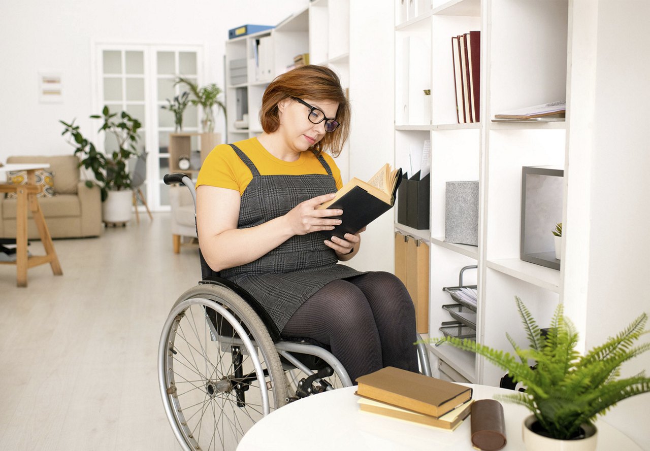 Female in a wheelchair reading a book and relaxing in a neutrally designed home, with soft, natural light illuminating the space and comfortable furnishings around her.