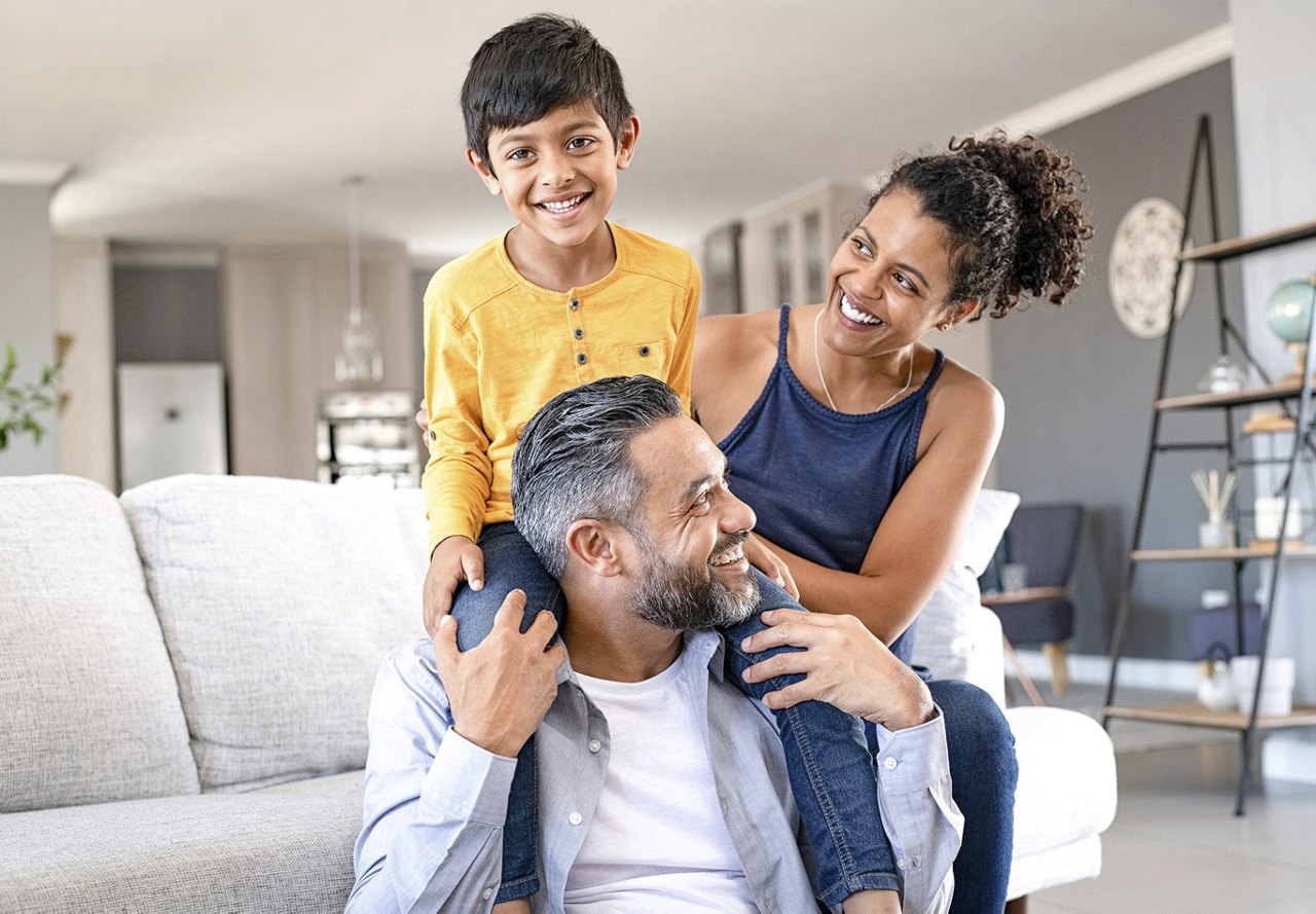 Family of three sitting together on the couch, smiling at each other and enjoying each other's company in a cozy home setting.