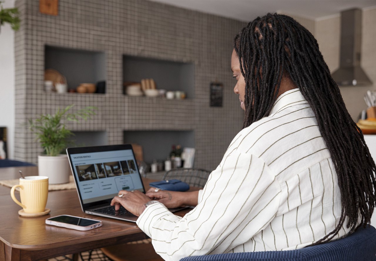 African American woman sitting at a table in the kitchen area, looking at apartment listings on her computer, with a phone and coffee next to her.