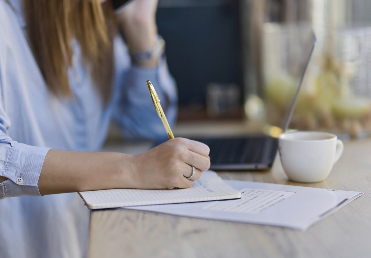 Woman in a blue button-up shirt on the phone, looking at a computer screen while writing notes on a notepad at a table, with a coffee mug and other sheets of paper scattered around.