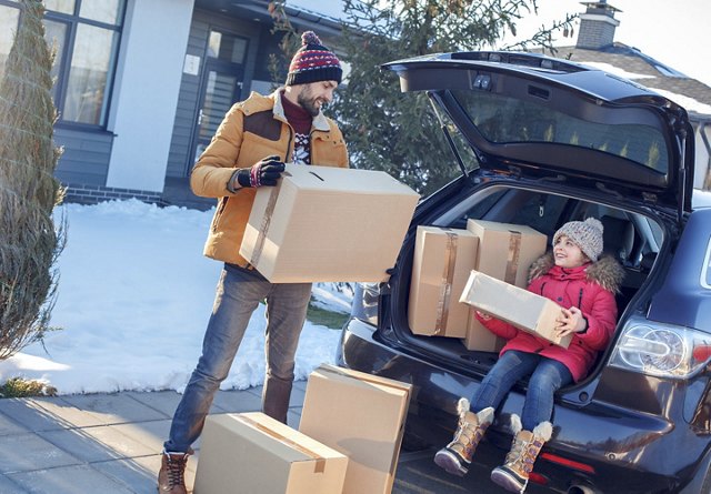 A father and daughter laughing together as they load their car with cardboard boxes for a move, set in a snowy winter scene with a Christmas tree nearby, both dressed warmly in coats and hats.