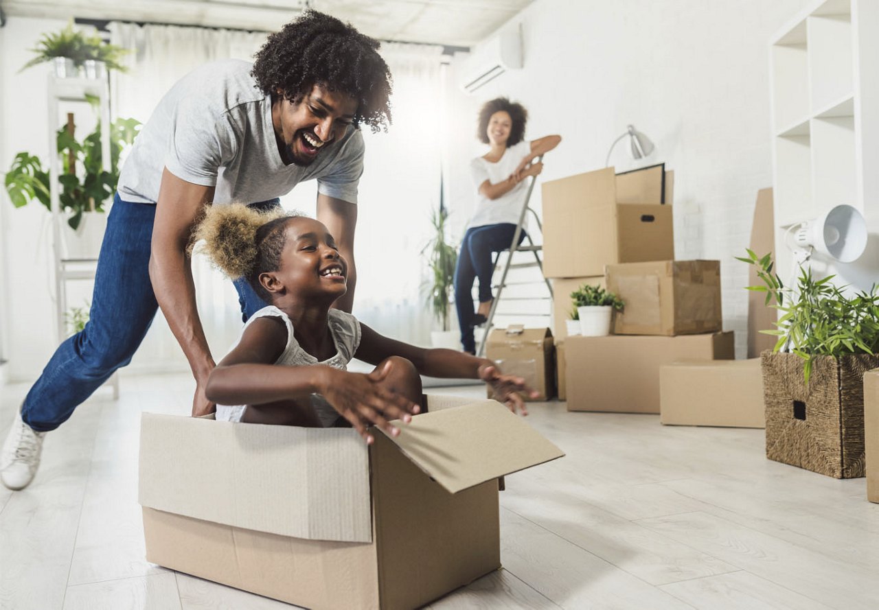 A family enjoying playful moments while packing for a move, with a father pushing his daughter in a cardboard box across the floor, the mother smiling as she watches, and packing boxes scattered around the room.