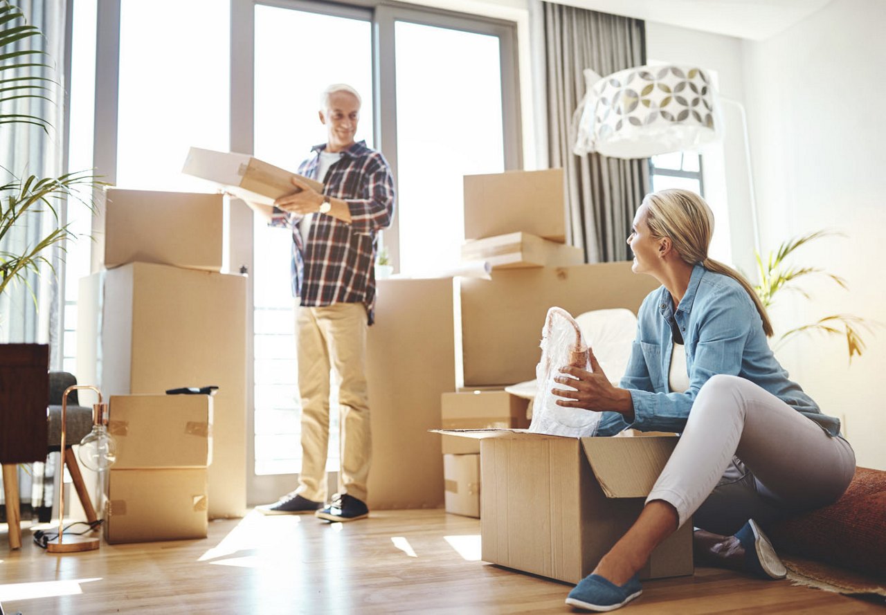 An older couple sharing a warm glance while packing boxes of their belongings in their home, surrounded by items being organized for their move, with sunlight streaming in through the windows.