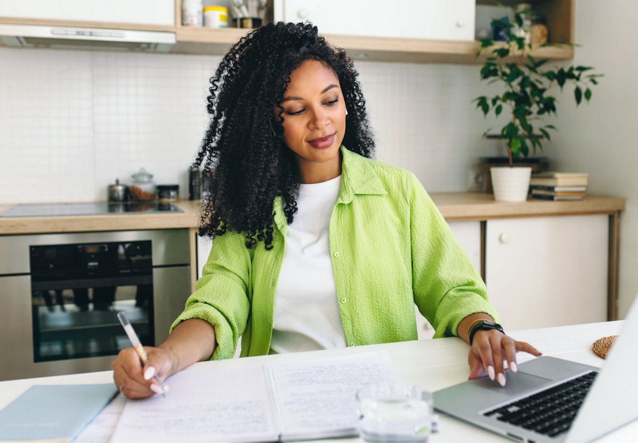 Woman sitting at a kitchen counter, looking at a computer and writing in a notebook, with the kitchen in the background as she creates a schedule.