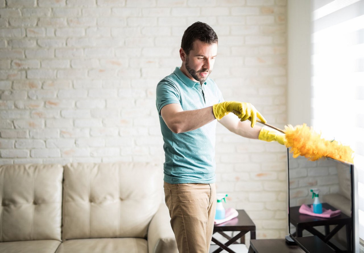 Man dusting a television in his apartment's living area, with an open brick wall and a couch in the background.