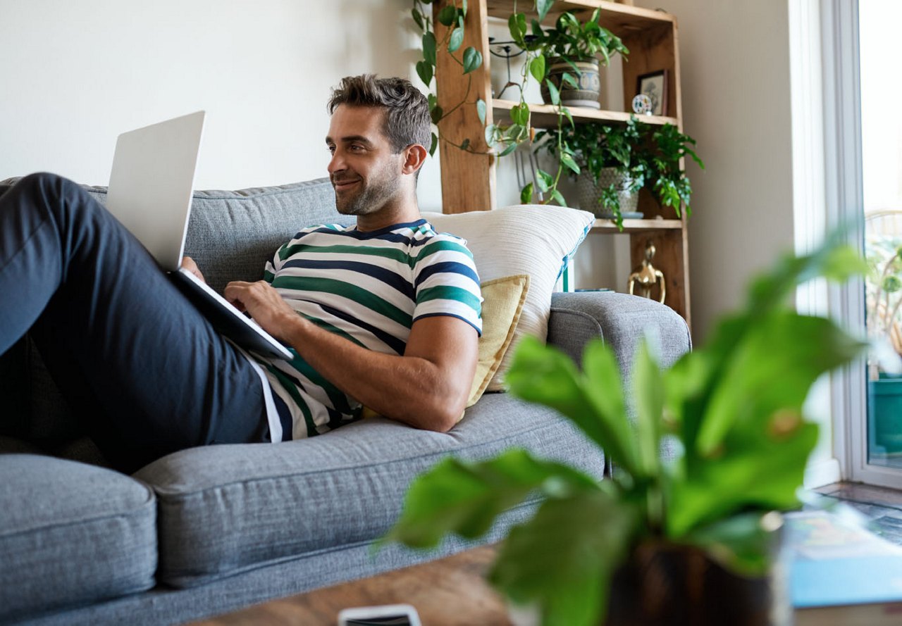 Man lying on the couch with a computer, smiling, surrounded by plants and sunlight illuminating his apartment.