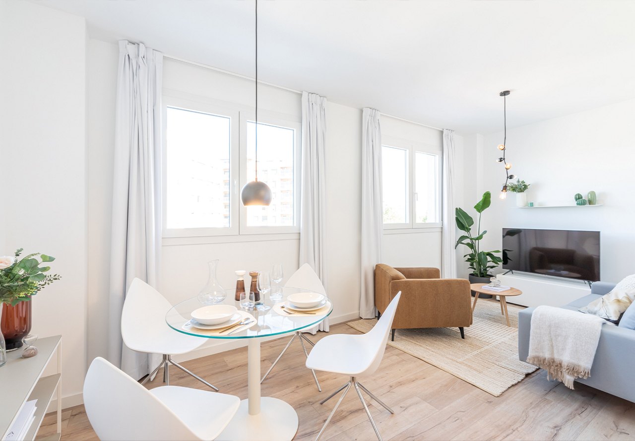 Modern apartment living space featuring a dining table, sitting area with a television, white walls, neutral decor, and touches of greenery.