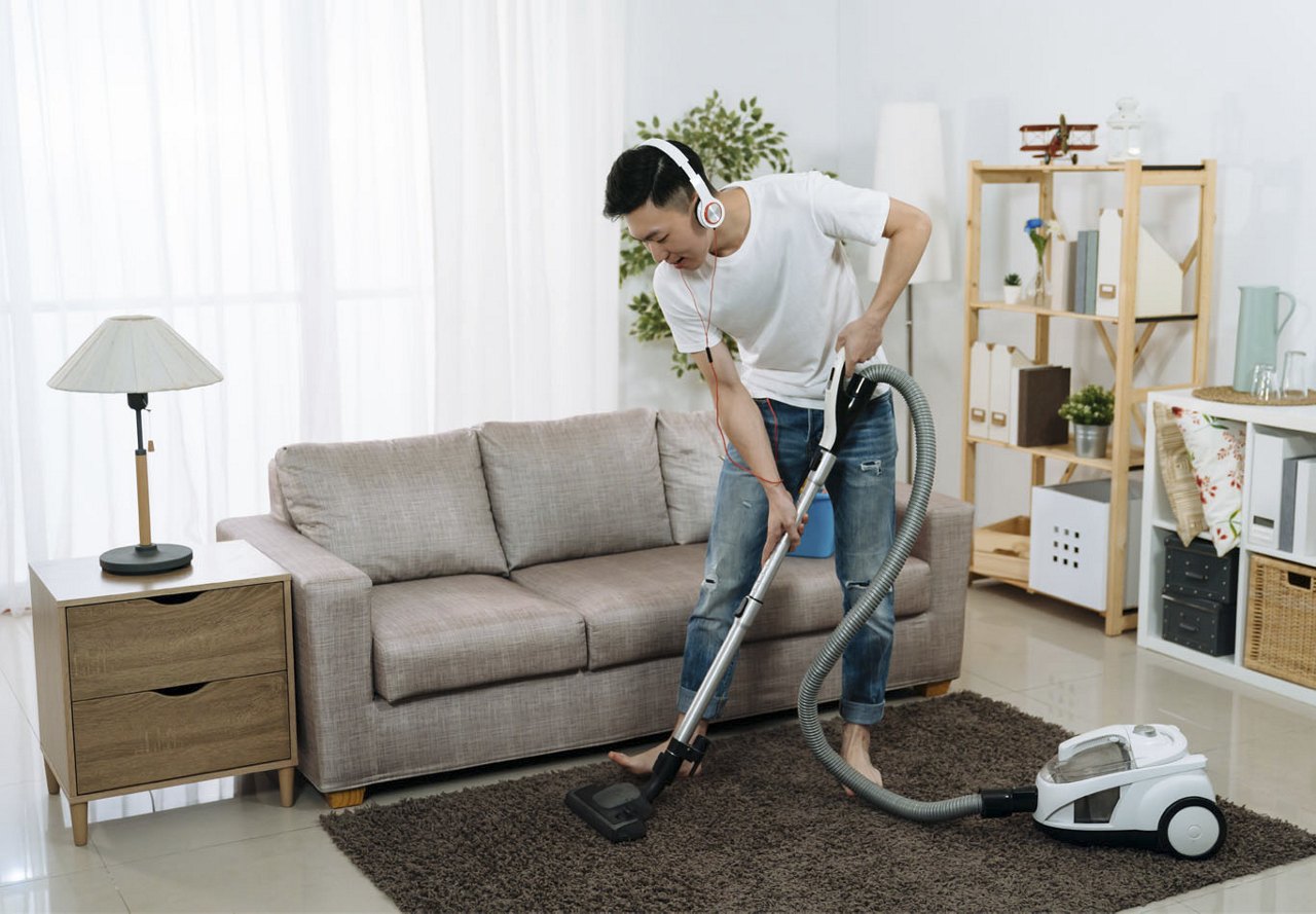 Man vacuuming the living room with headphones on, cleaning a rug while surrounded by stylish home decor.