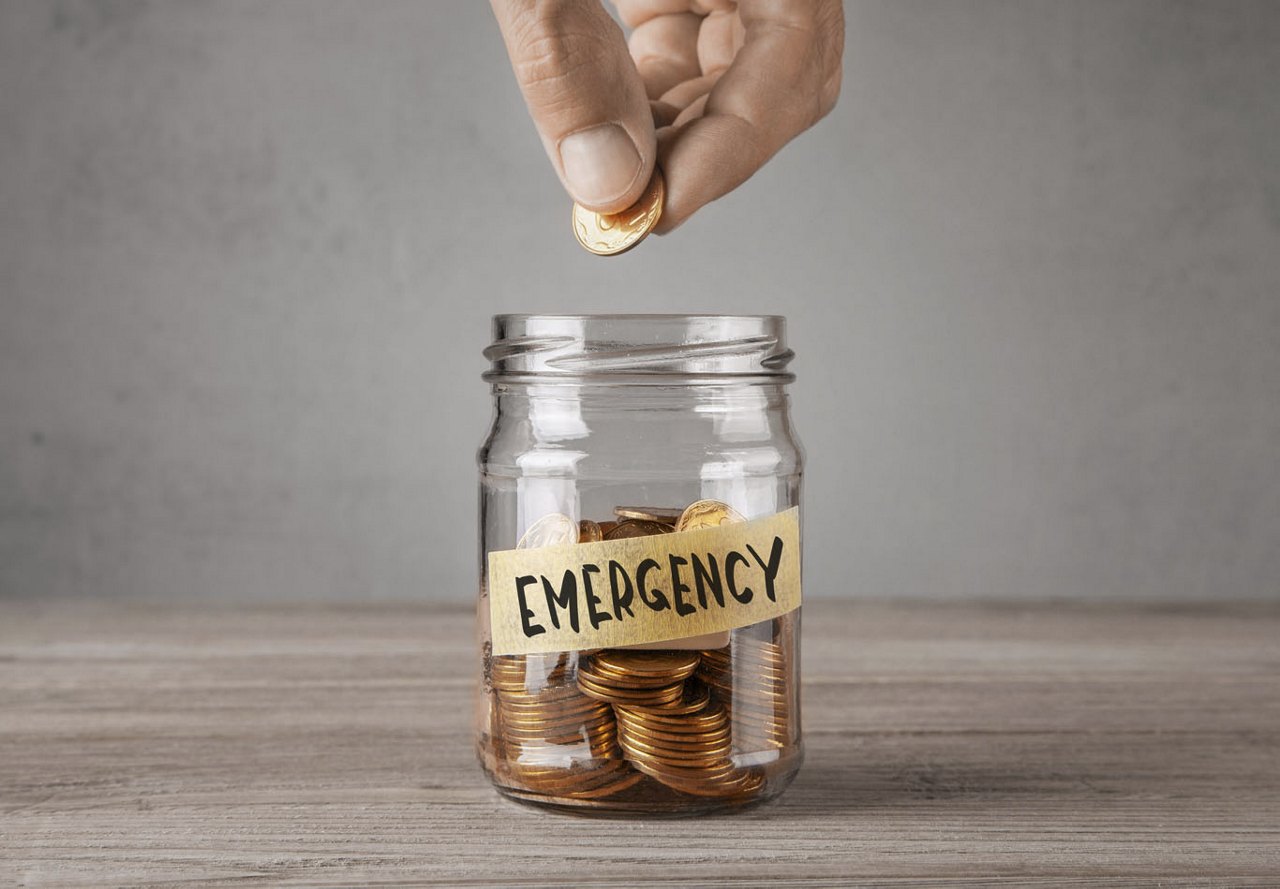 Hand placing a coin into a glass jar labeled 'Emergency' for savings designated for emergency purposes.