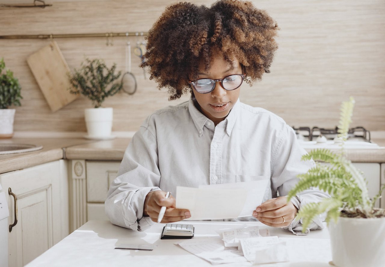 African American woman sitting at her kitchen table reviewing financial documents with a calculator and pen, surrounded by household plants.