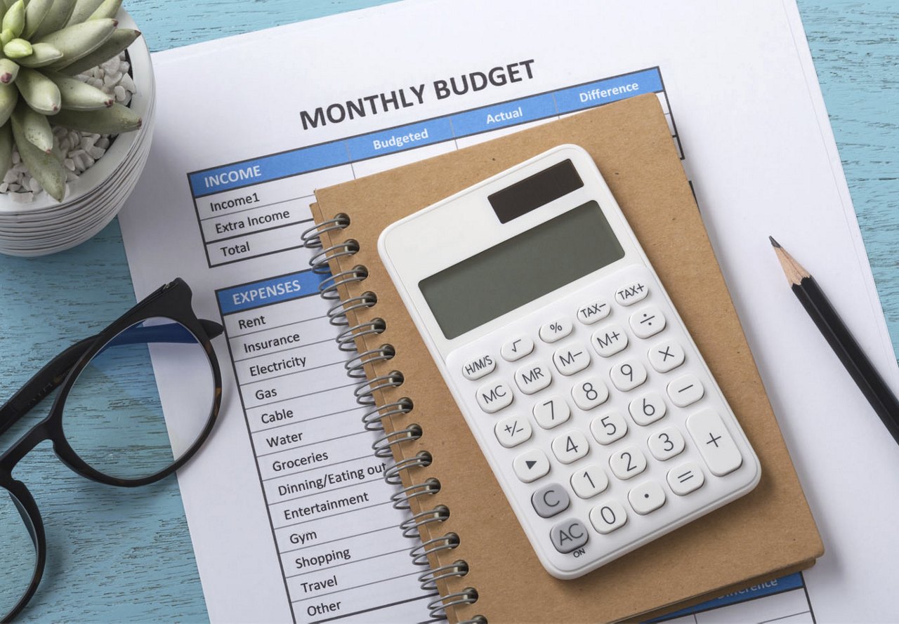 Blue wooden table with a printed monthly budget on paper, a small brown notebook, calculator, glasses, and a small plant arranged neatly.