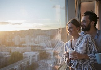 A couple standing by their apartment window, smiling as they hold cups of coffee, enjoying the view of the city below and preparing for the day ahead.