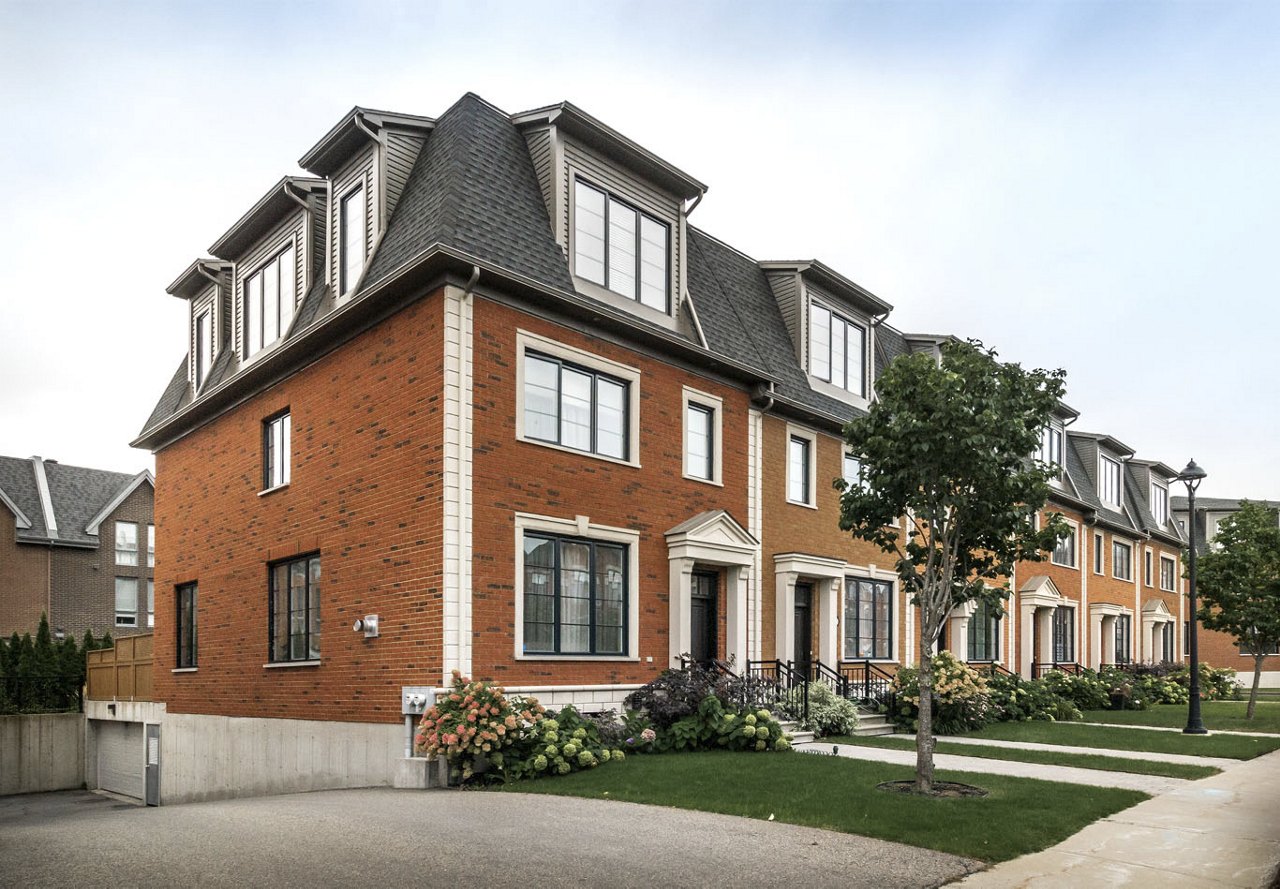 A street view of a charming brick townhome with a tree in the front yard, vibrant flowers near the entrance, and a welcoming atmosphere, set against a clear blue sky with a sidewalk leading up to the home.