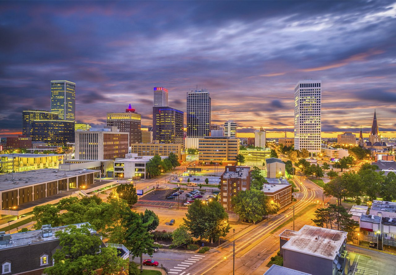 A view of Tulsa, Oklahoma, with vibrant city lights illuminating the skyline, set against a beautifully colored sky as day transitions into night.