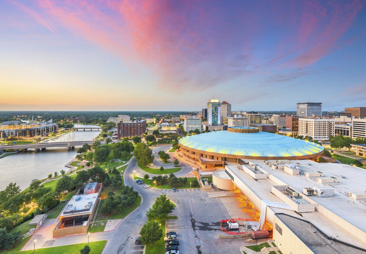 A view of Wichita, Kansas, with a stunning pink and blue sunset, showcasing the city below, surrounded by lush trees and a river on the left side.