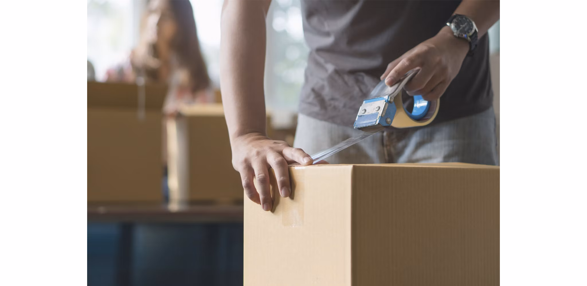 Man taping up a box while preparing for a move, with a woman in the background also helping with the packing process.