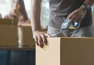 Man taping up a box while preparing for a move, with a woman in the background also helping with the packing process.