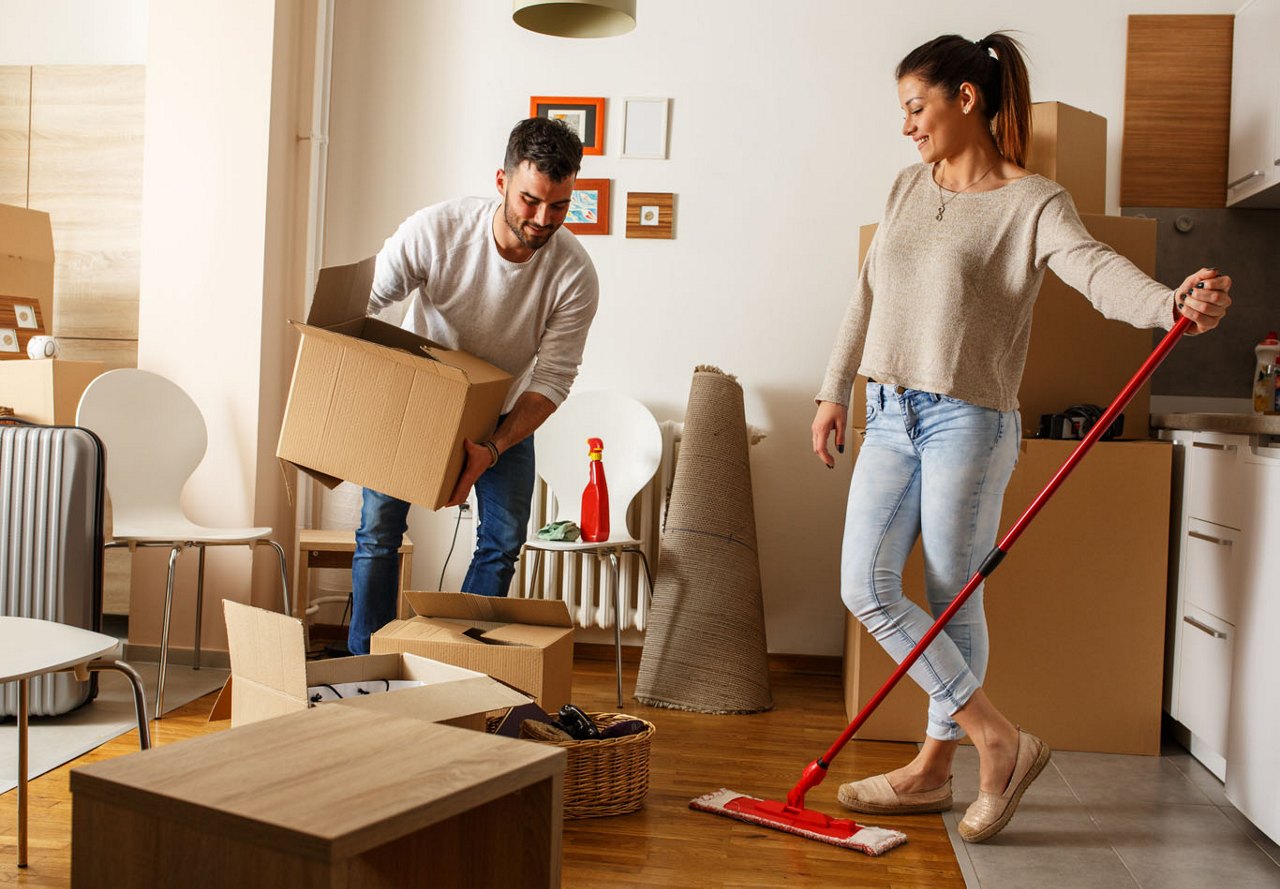 Couple cleaning their apartment together while moving out, with boxes around them and the woman holding a mop, preparing the space for departure.