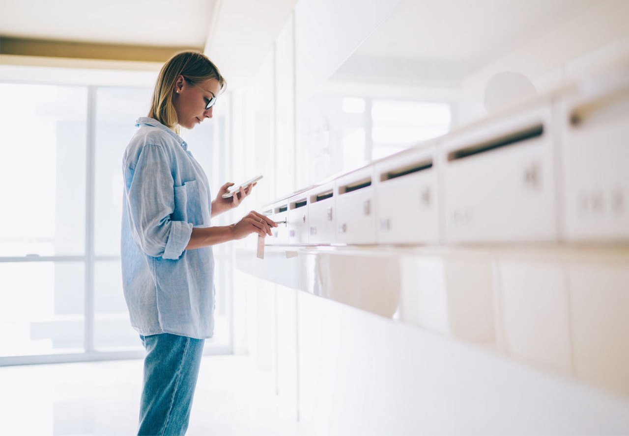 Woman standing at mailboxes in an apartment complex, holding her phone and carefully inserting a key to open her mailbox, with a blurred mailroom in the foreground.