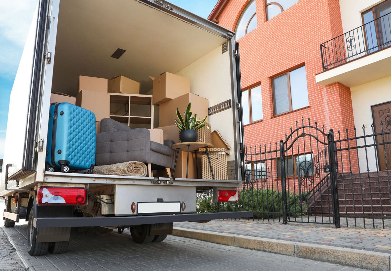 Moving truck with the back open, as people load items and furniture while moving out of an apartment, with apartment buildings visible to the right.