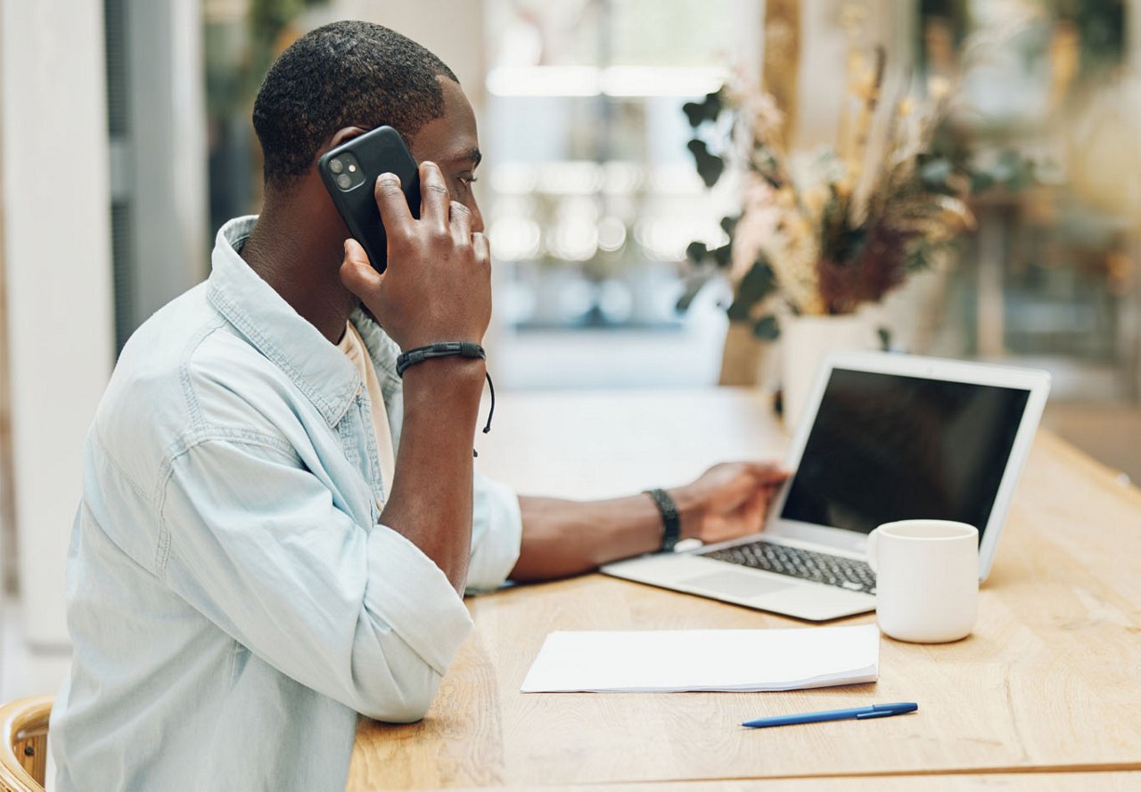 African American man sitting at his computer, talking on the phone while looking at the screen, with a coffee mug and supplies on the table as he works.