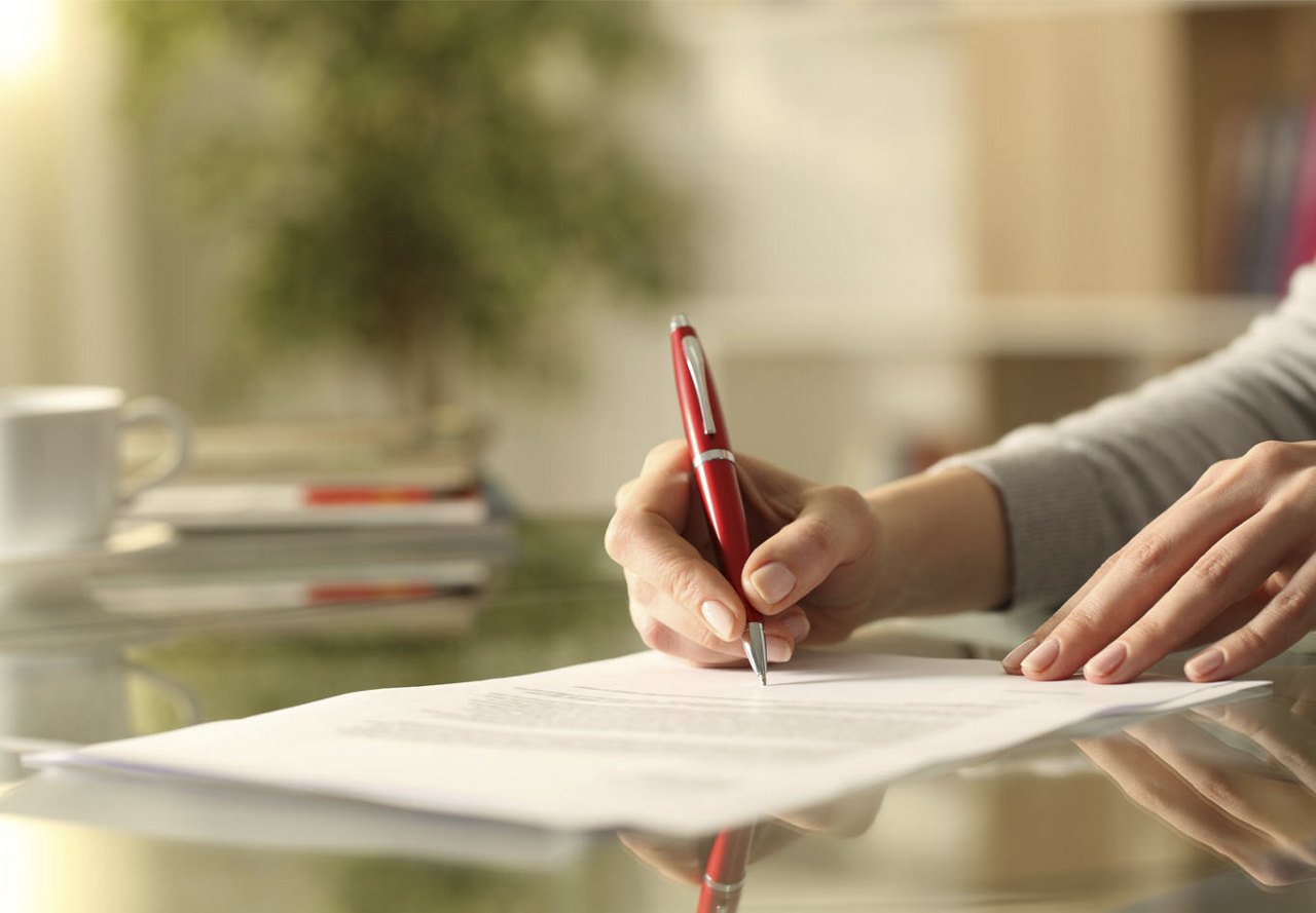 Person sitting at a desk, writing on a piece of paper with a red pen, signing a document, with a blurred room in the background.