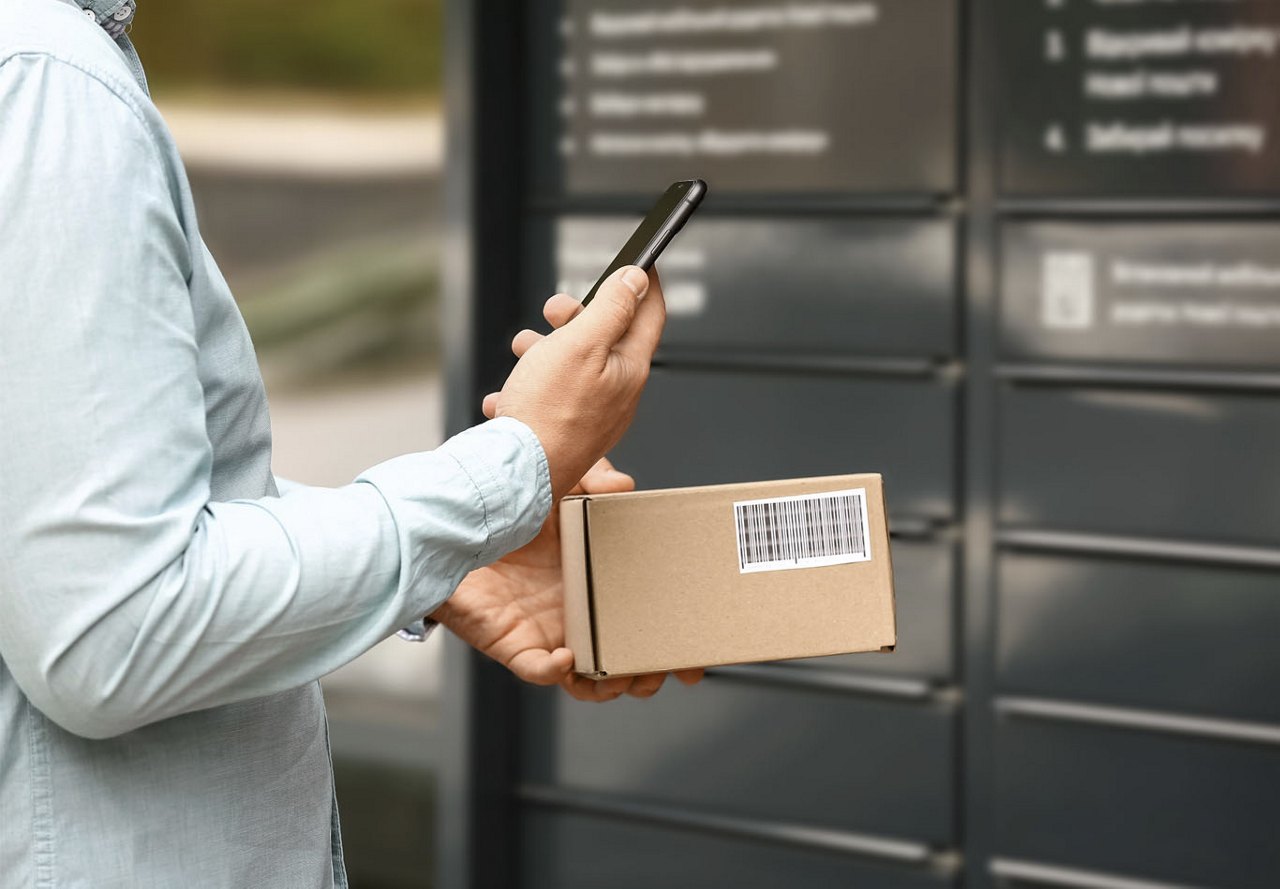 A person holding a cardboard package and looking at their cell phone after retrieving it from a storage locker, with the locker blurred in the background.