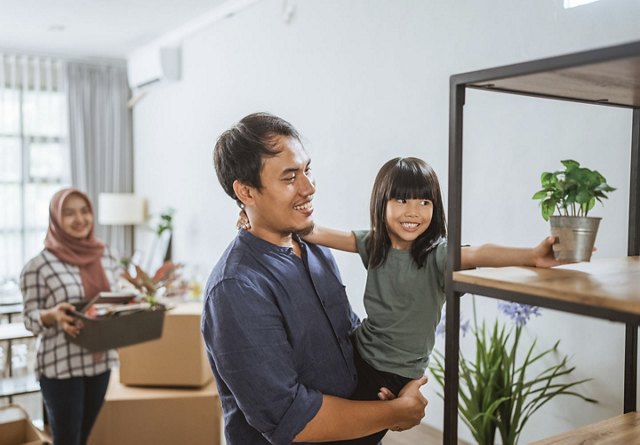 Family settling into their new home, with the father and daughter placing a plant on a shelf, while the mother watches them smiling in the background.