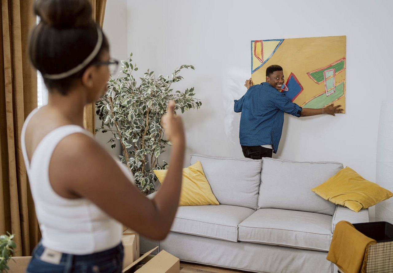 Couple hanging wall art in their home, with the man holding the art up to show its placement while the woman guides him on where to hang it.