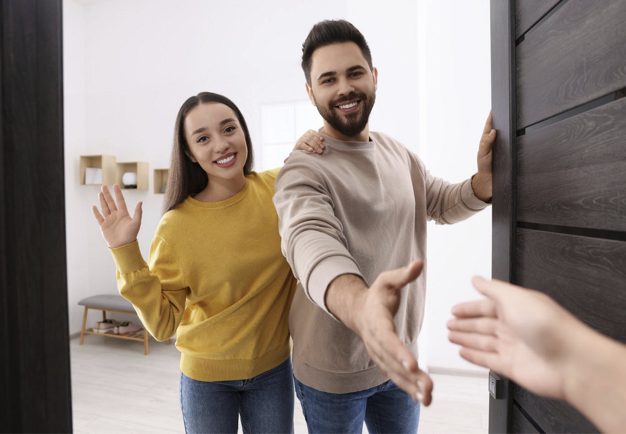 Couple greeting a neighbor at their apartment door, with the man reaching out for a handshake and the woman waving.