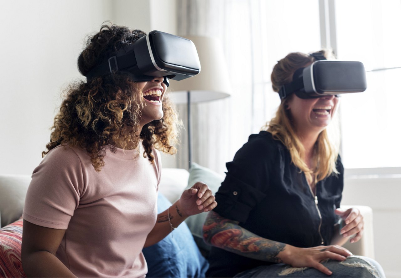 Two women laughing and enjoying a virtual reality experience, both wearing VR headsets, seated on a couch.