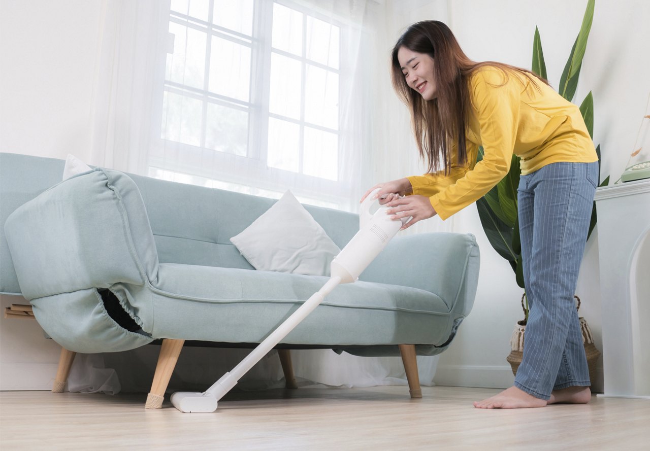 A smiling woman in a yellow shirt uses a handheld vacuum cleaner on a light blue sofa in a bright living room