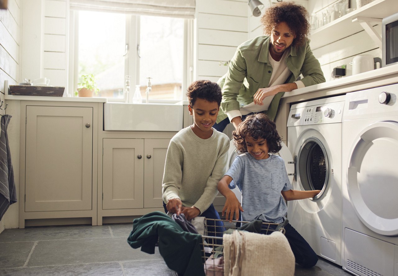 A father and two children happily do laundry together in a sunny home interior.