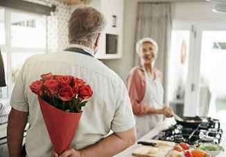 A man surprises a woman in the kitchen with a bouquet of red roses hidden behind his back.