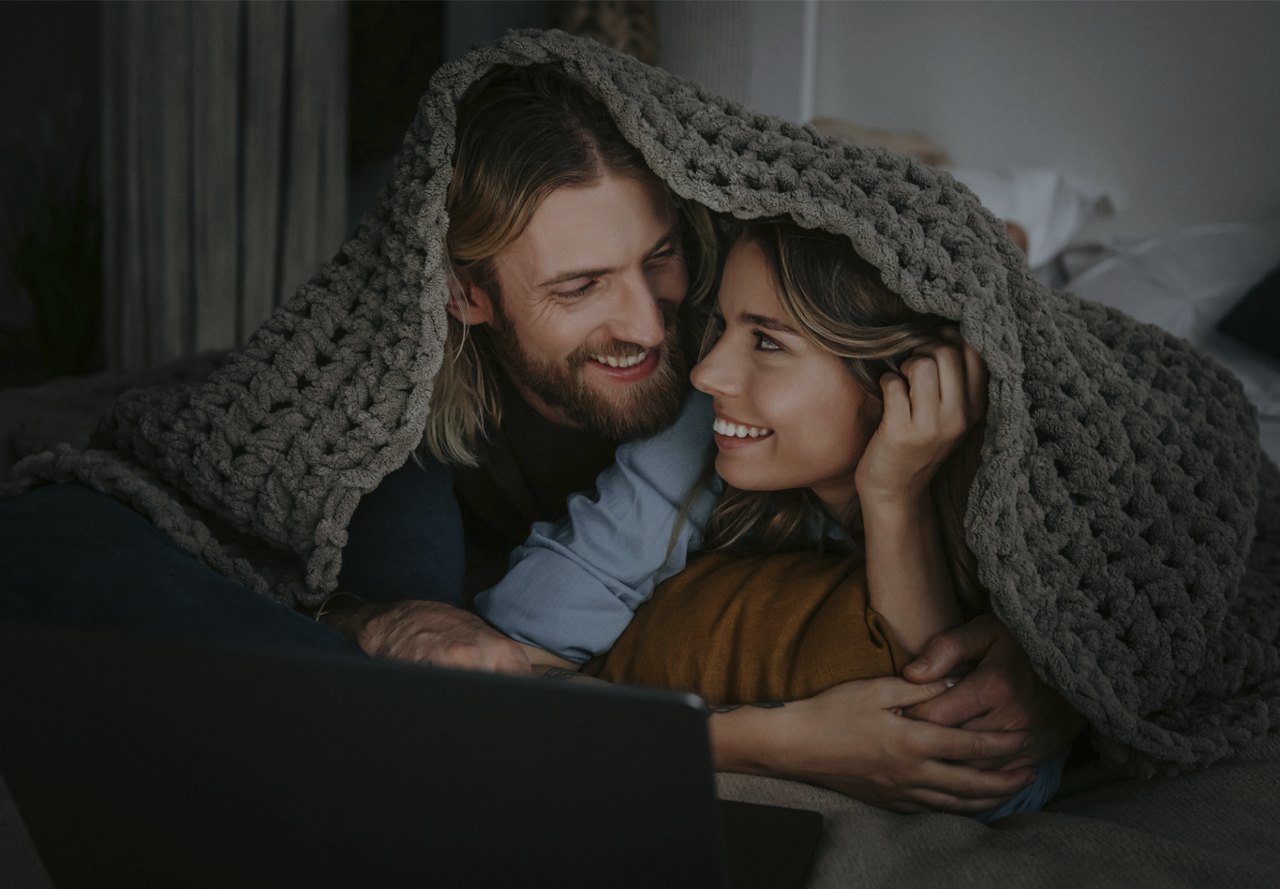 A couple shares a warm embrace under a cozy blanket while looking at a laptop screen.