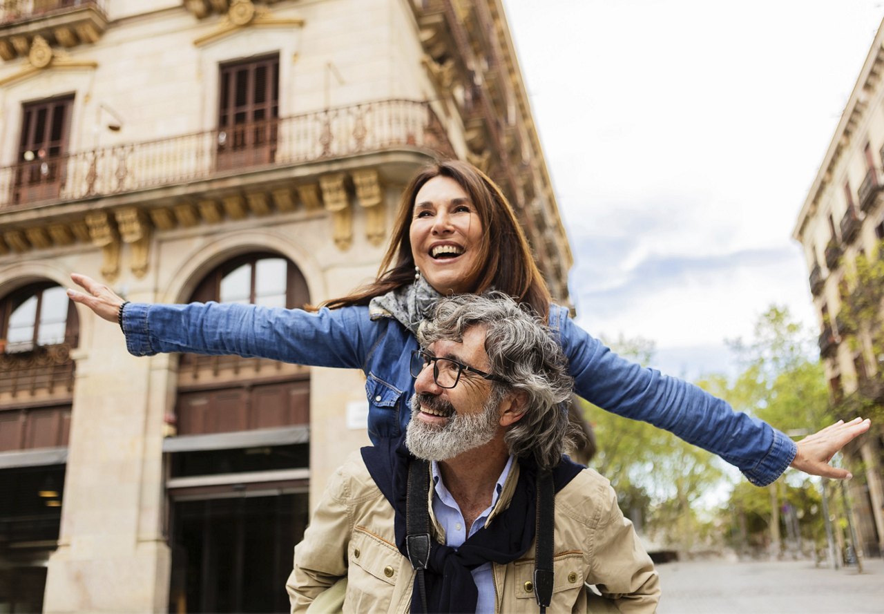 A joyful woman with outstretched arms rides piggyback on a smiling man in an urban setting.