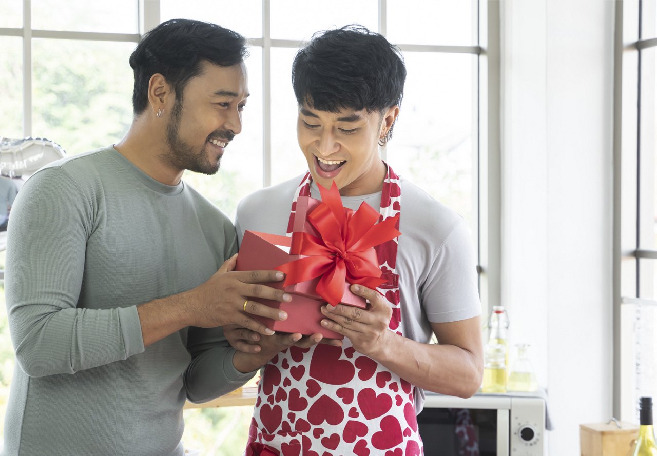 One man surprises another with a red gift box, both smiling in a bright kitchen.