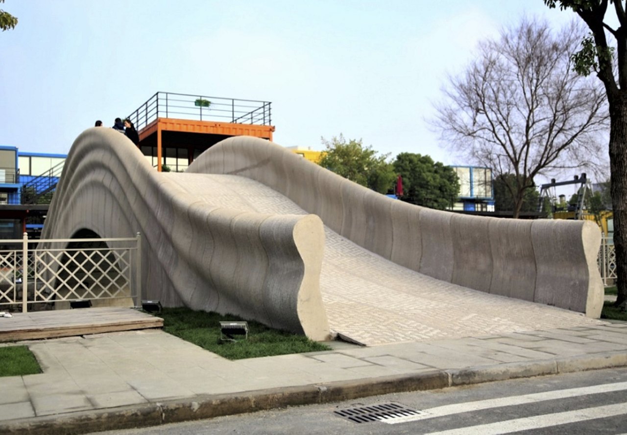 A unique 3D-printed pedestrian bridge with wavy contours and textured surfaces in an urban setting.